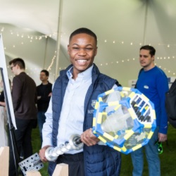 A person smiling broadly holds a large, colorful geometric object with clear panels in an event tent. Other attendees are visible in the background, engaged in activities 和 discussions. The tent is decorated with string lights.