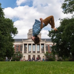 A person is mid-air performing a backflip on a grassy field with a large, stately building with columns 和 a dome in the background. The sky is partly cloudy 和 there are several people 和 trees in the vicinity.